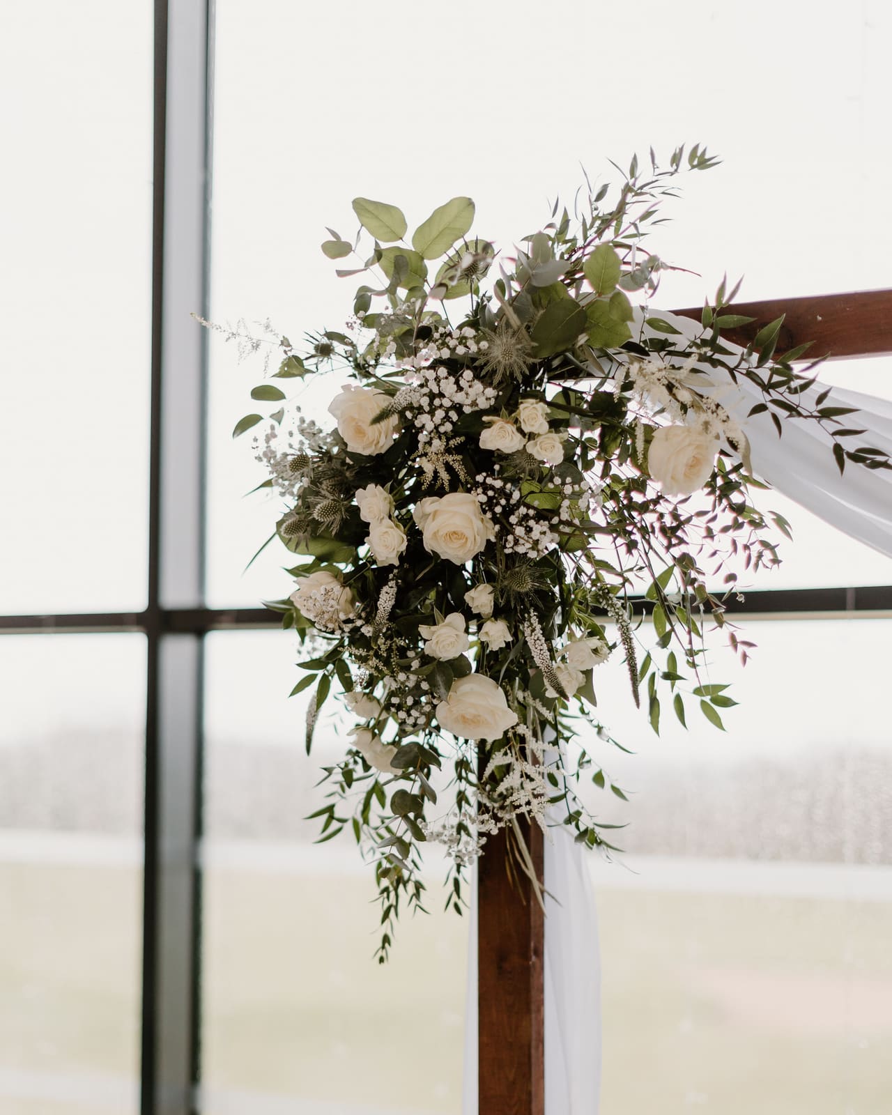 White roses and eucalyptus on a timber ceremony arch beside a tall window over open fields.